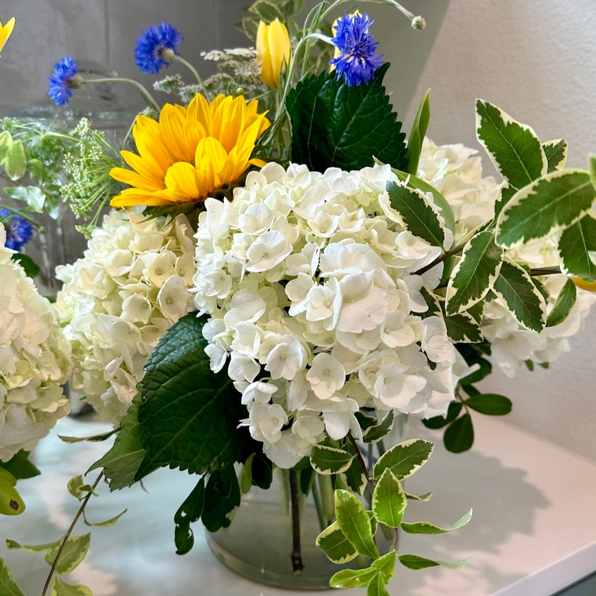 Floral arrangement with white hydrangeas, yellow sunflowers, and greenery on a light surface.